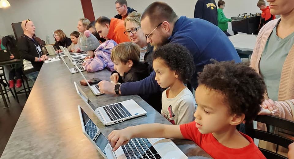 Elementary-aged students gather around computers at NewBoCo while a volunteer teaches them about programming.