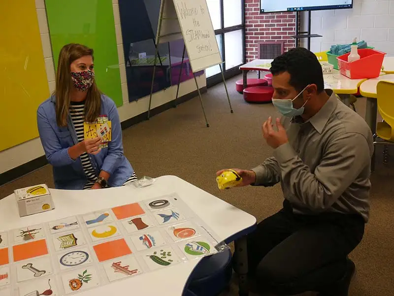 Fort Madison Community School District teachers chat and work together in a CSforALL robotics programming exercise that places them around a table with a grid of images that they must navigate by programming a fist-sized, bee-shaped robot.