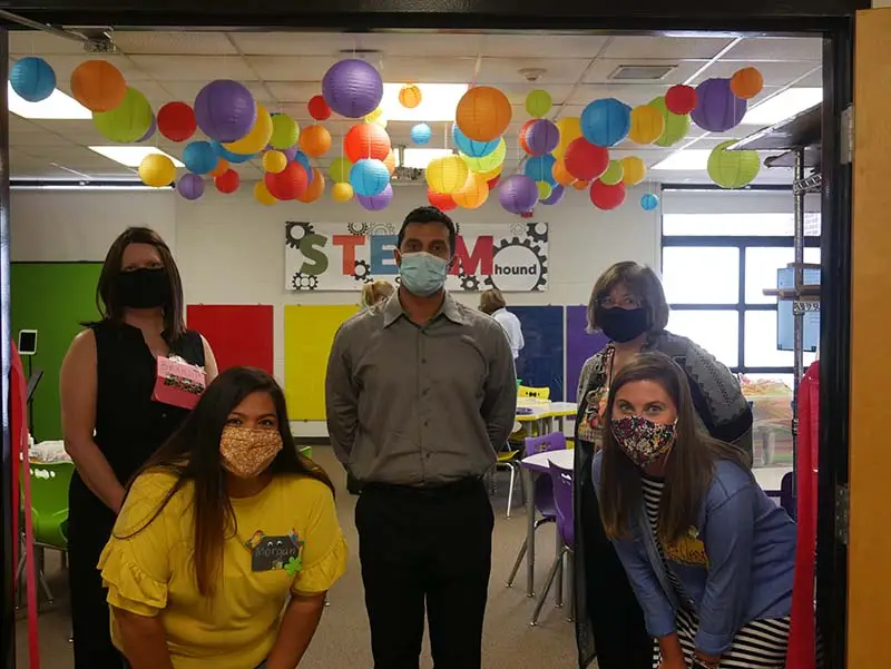 5 people in various positions and sanitary masks in front of a brightly decorated school room with hanging paper lanterns.