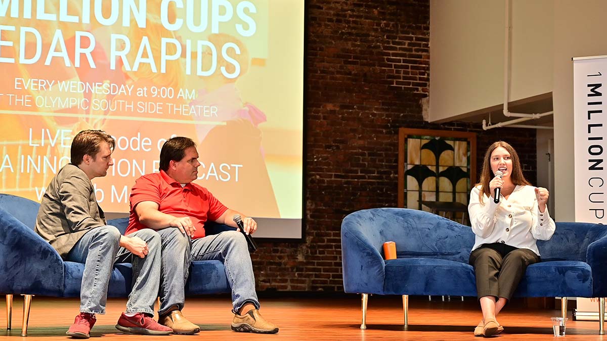 2 hosts sit on a couch on the 1 Million Cups stage while their interviewee speaks into a microphone from a couch beside them