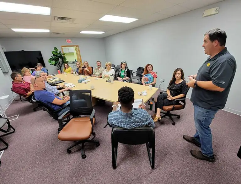 15 people sit around a conference table and listen attentively to 1 person who stands before them and gesticulates during the presentation
