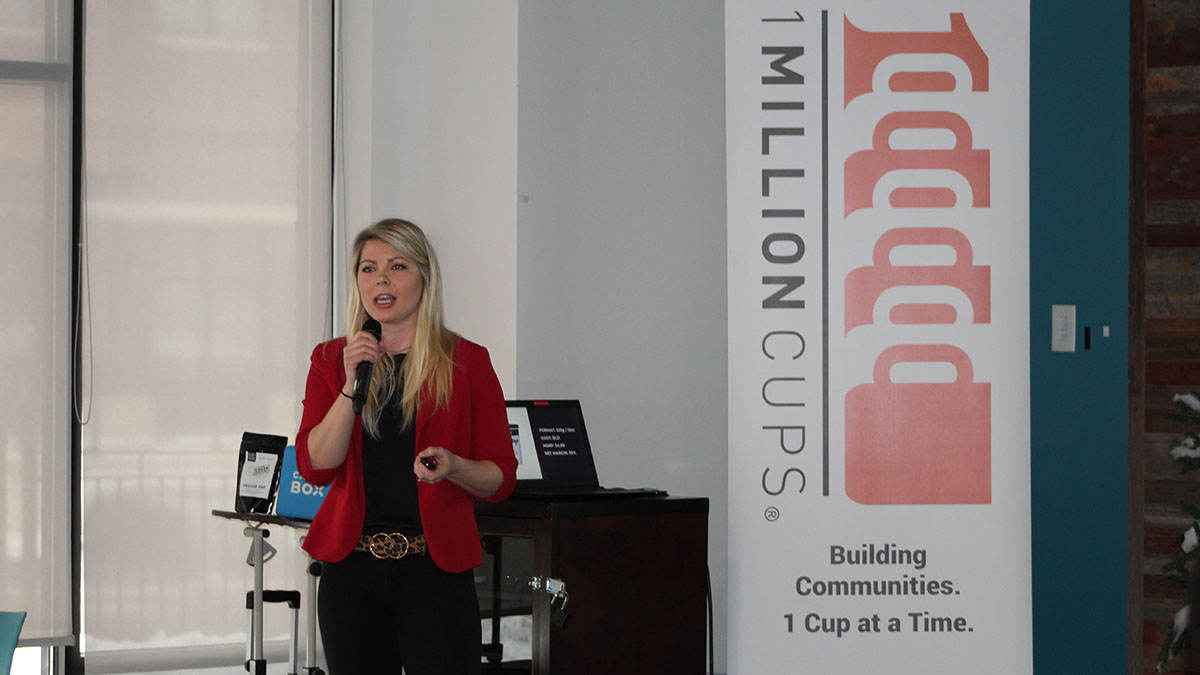 A small business owner presents to an audience from in front of a 1 Million Cups banner