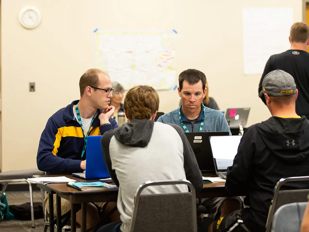 3 Code.org teachers stare intently at their laptops while another teacher speaks to them