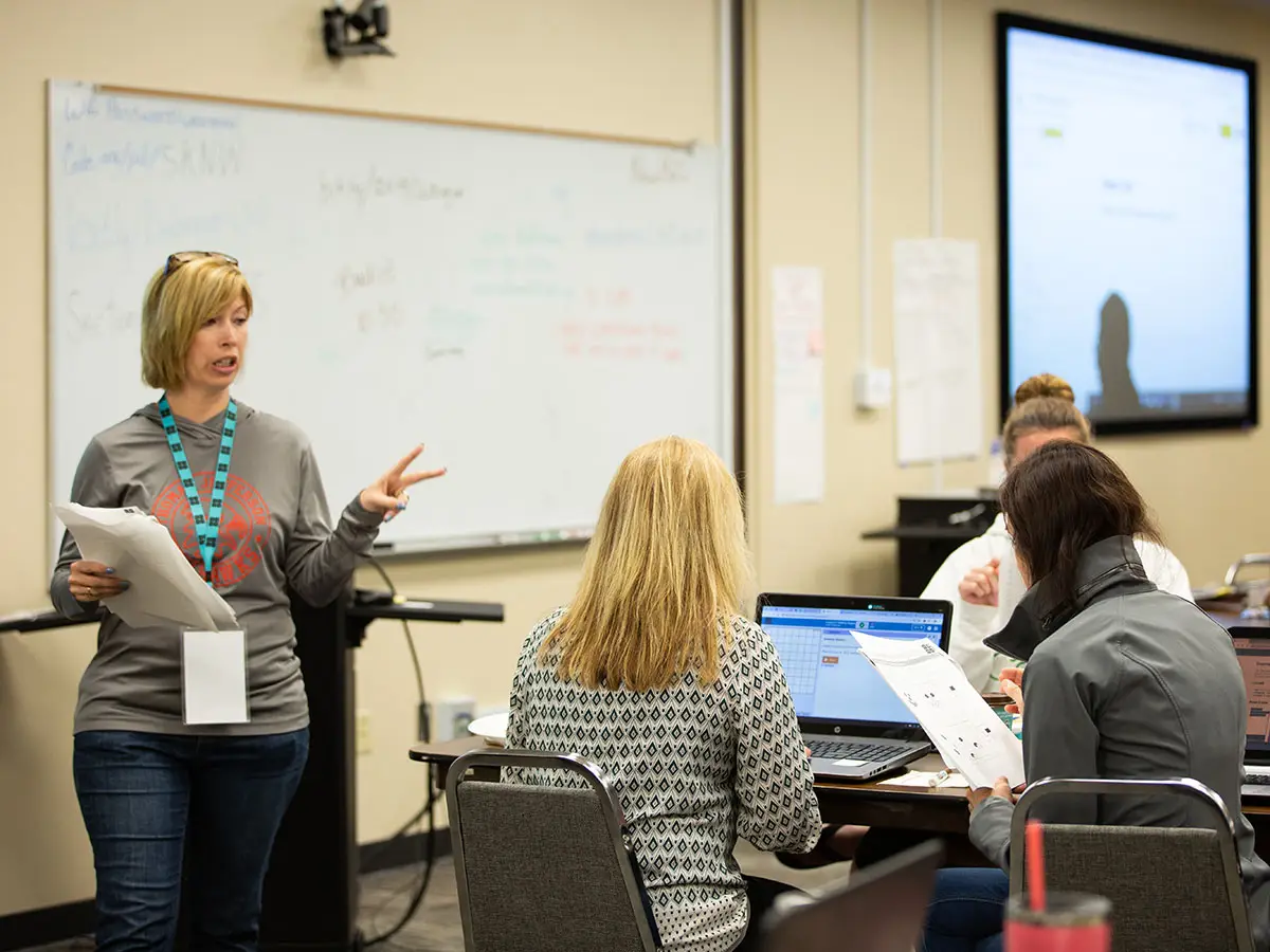 A Code.org teacher forms the number "2" with her fingers as she speaks with 3 other seated teachers