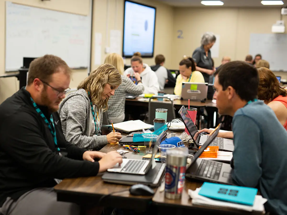 A classroom full of Code.org teachers work studiously at their notebooks and laptops as a facilitator stands amongst them