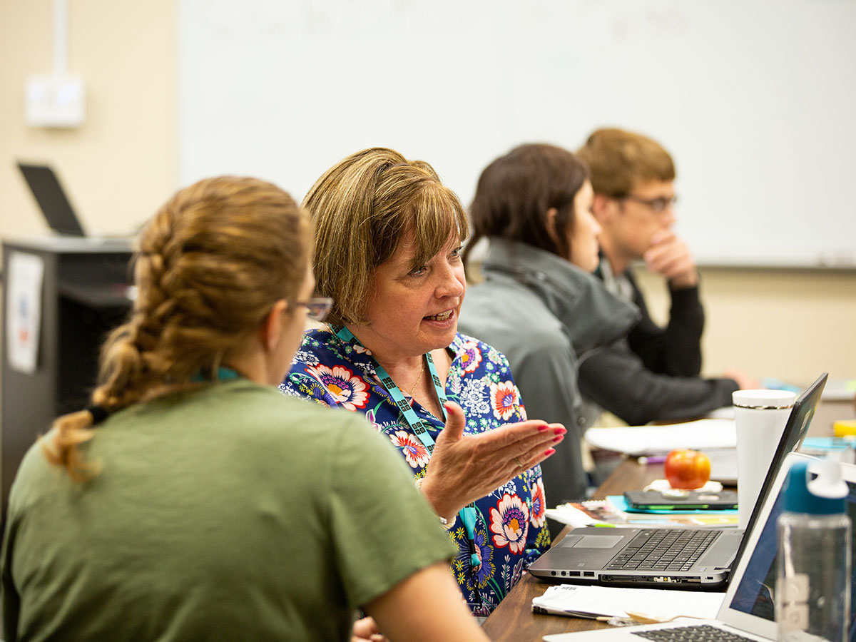 A teacher speaks and gesticulates as another group member listens intently; another group studies a laptop screen in the background