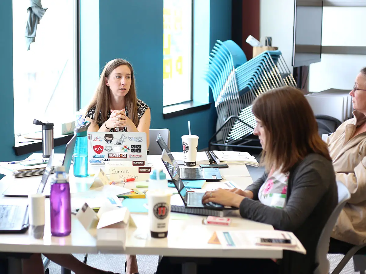 A Code.org facilitator sits at a table and speaks with 4 teachers; laptops and notebooks are strewn about the table