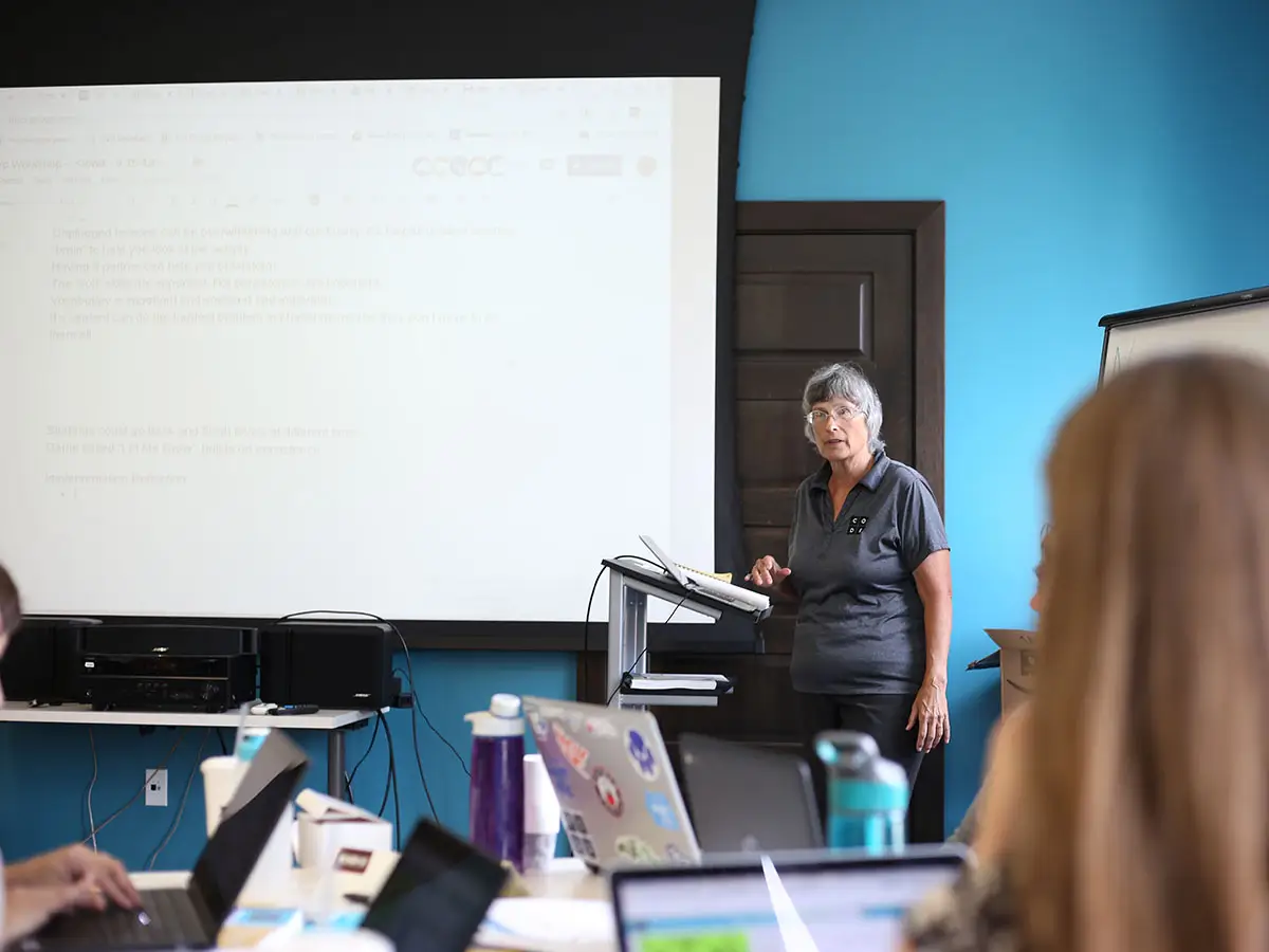 A Code.org facilitator stands at a podium in front of a projection screen and speaks to a class