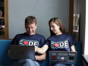 2 NewBoCo employees site behind their laptops and showcase their "Code" t-shirts with hearts replacing the "O's"