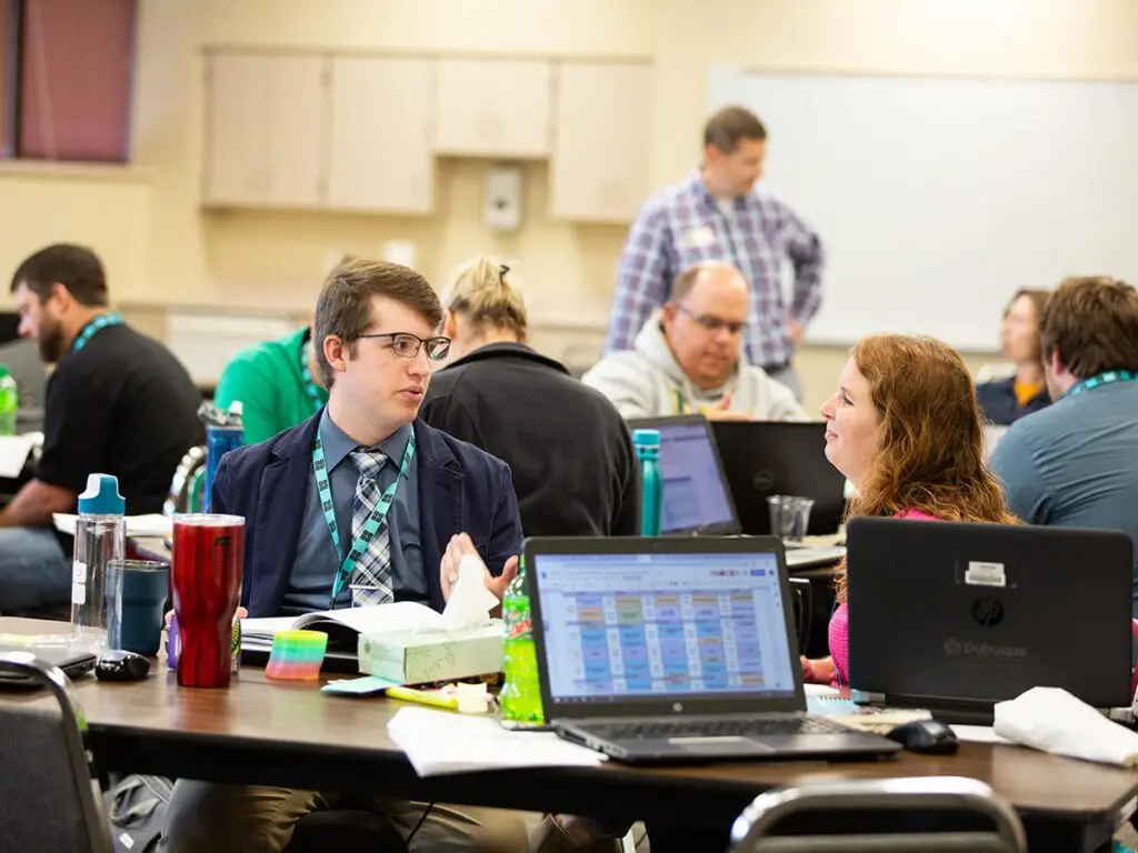 2 seated Code.org teachers hold a conversation, while more teachers in the background work with their heads down