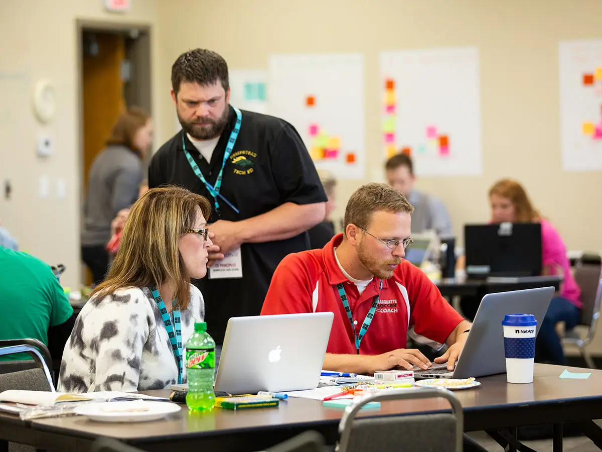 A Code.org facilitator looks over the shoulder of 2 teachers as they work together at a laptop