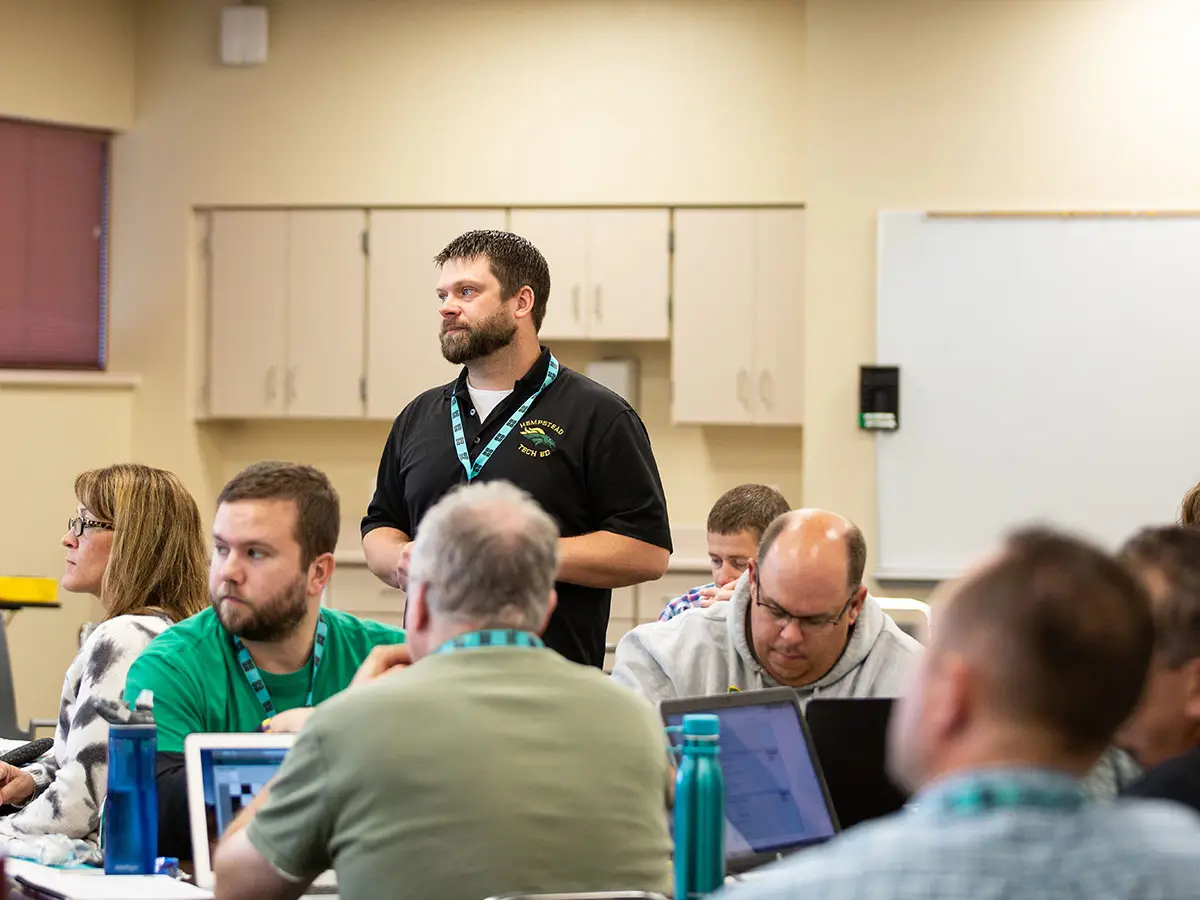 1 Code.org facilitator stands and 7 students sit at tables, all looking out of frame with serious expressions