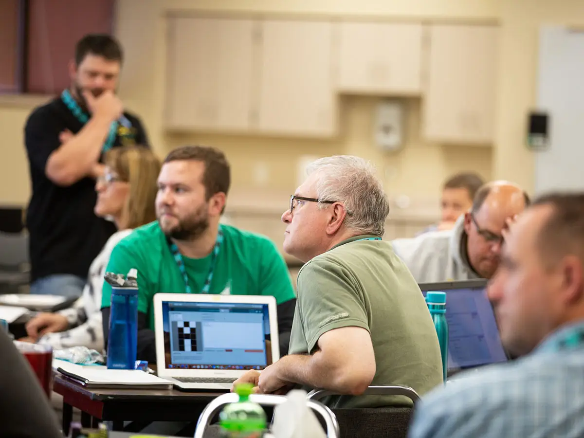 5 Code.org teachers gaze out of frame in the foreground, while in the background a facilitator stands and scratches his chin, contemplating something on a table