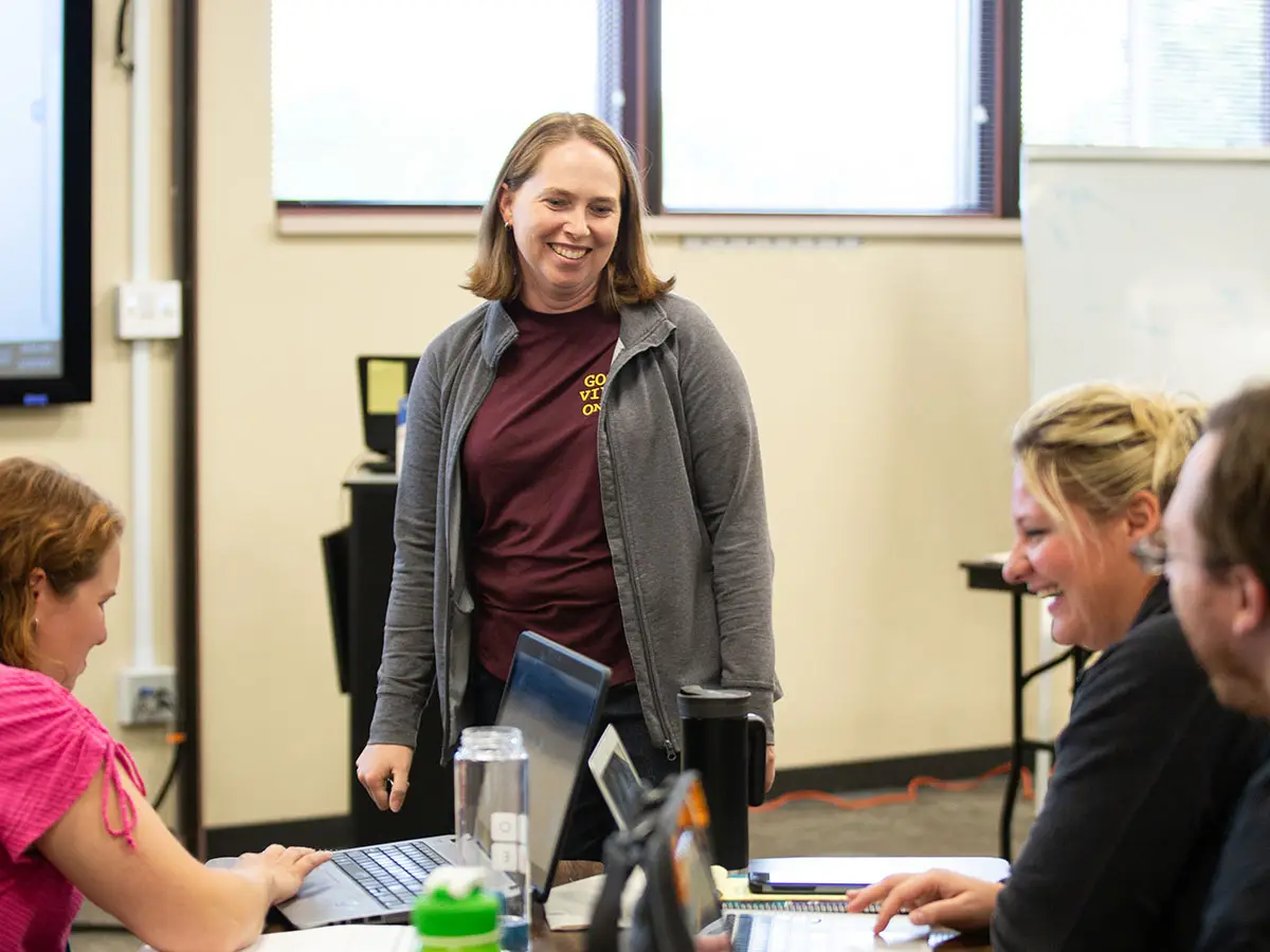 1 teacher stands and speaks with 3 other group members who sit at a table; they are all laughing