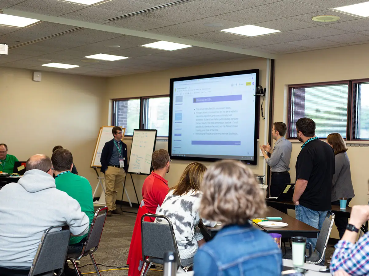 A classroom full of Code.org teachers look on at a projector slide show primarily of text while a facilitator speaks