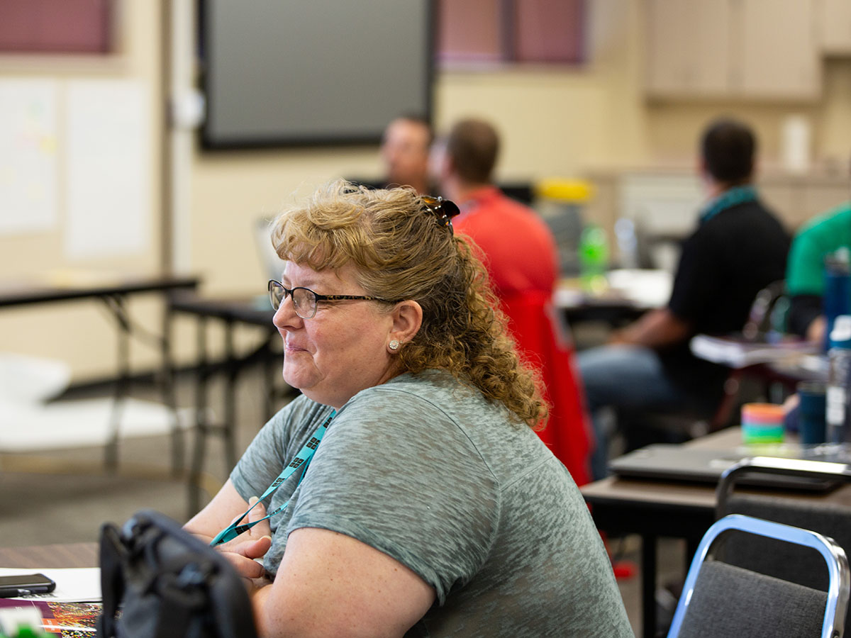 A teacher sits at a table and smiles; another group sits out of focus in the background