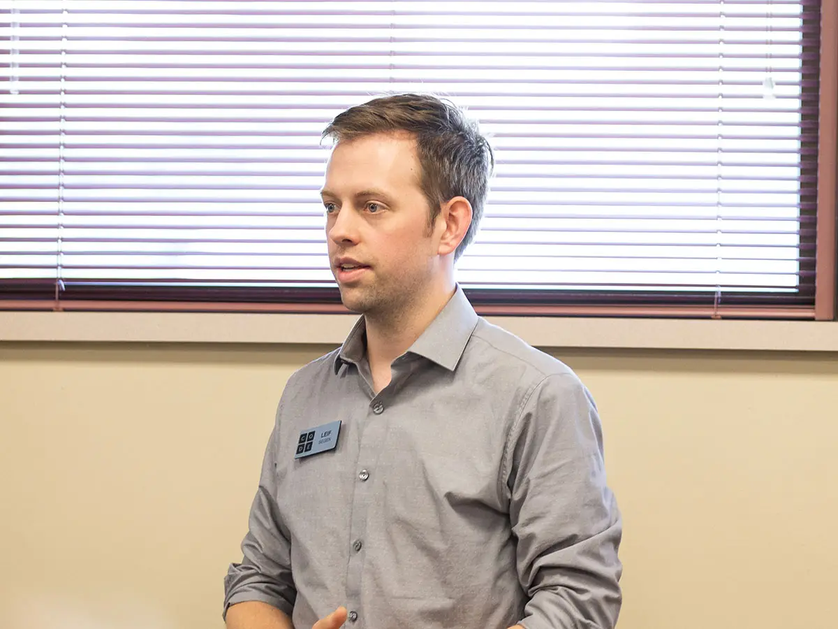 A code.org facilitator speaks from in front of a pale yellow wall and closed window blinds