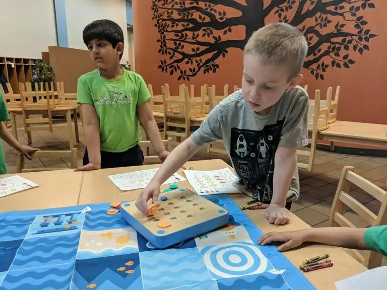 A Tiny Techies student places pegs into a wooden imitation circuit board while another student looks out of frame