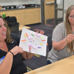 2 Tiny Techies teachers smile and show off projects they made by drawing on and pasting different items to a paper