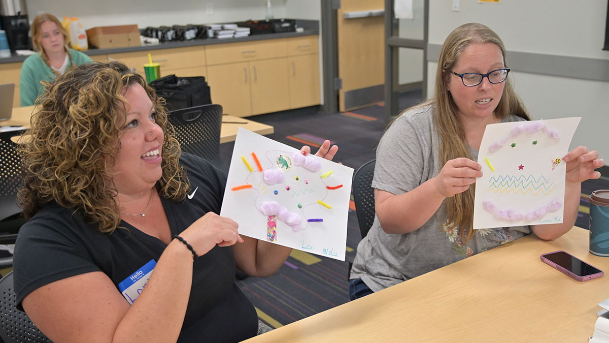 2 Tiny Techies teachers smile and show off projects they made by drawing on and pasting different items to a paper