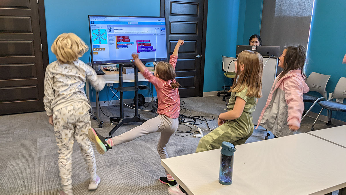 Girls Who Code Club participants dance in front of a TV displaying blocks of code the participants assembled.