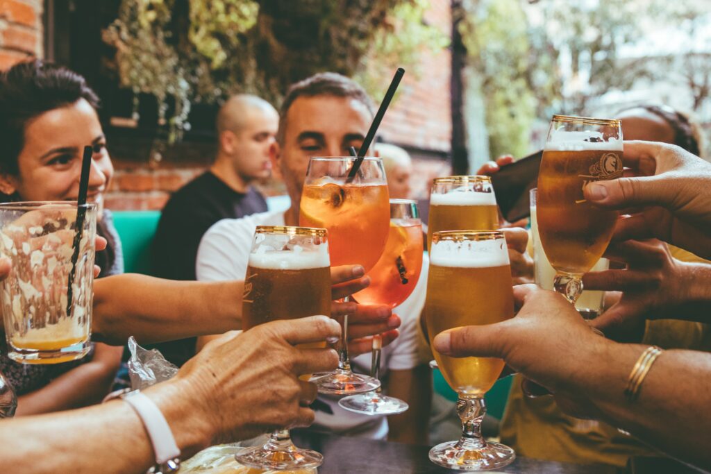 A group of coworkers surround eachother holding a cocktail or spirit upright and clinking their glasses. They are sitting inside a restaurant .