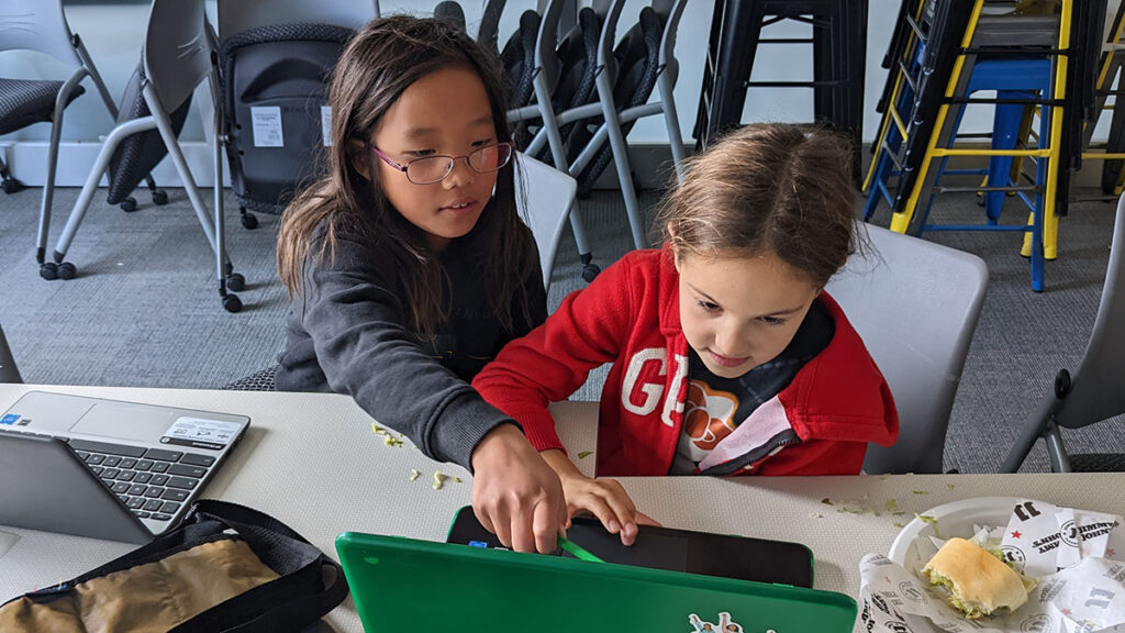 A young Girls Who Code Club participant points at her classmates laptop screen to help her