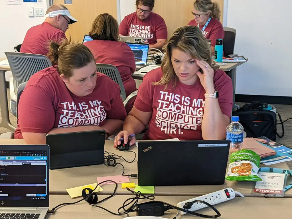 In the foreground, 2 Code.org workshop participants stare exasperatedly at a computer screen; in the background, 4 more around a table look at their laptops in front of them