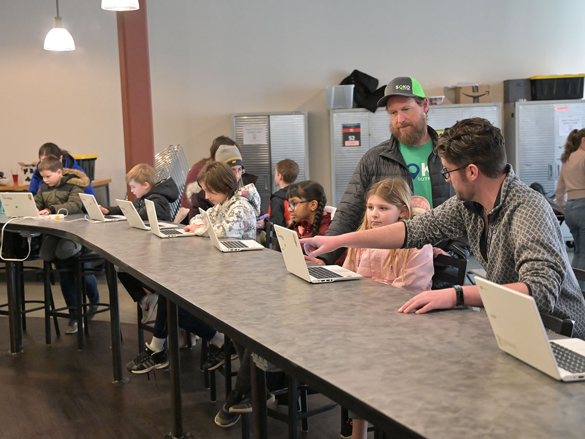 A CoderDojo facilitator and volunteer help a participant through an exercise; they point at a laptop screen a long a row of more children working at laptops