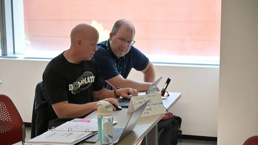 2 Project GUTS participants sit at a table and work together; 1 looks at the viewer
