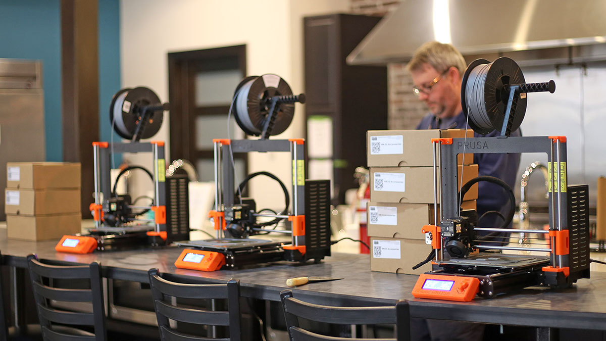 In the foreground is a row of 3D printers on a counter; in the background, a NewBoCo employee looks down, busy with a task