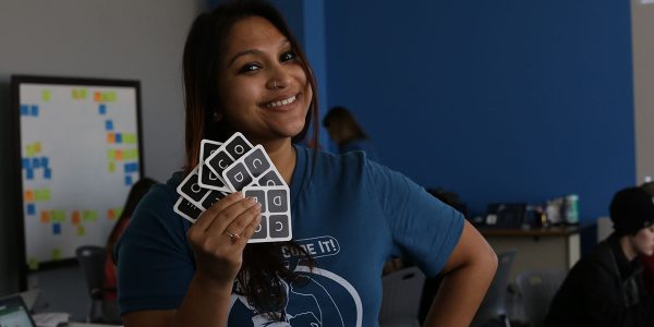 A smiling person stands, hand on hip, and holds up a fanned series of cards with "C-O-D-E" on them