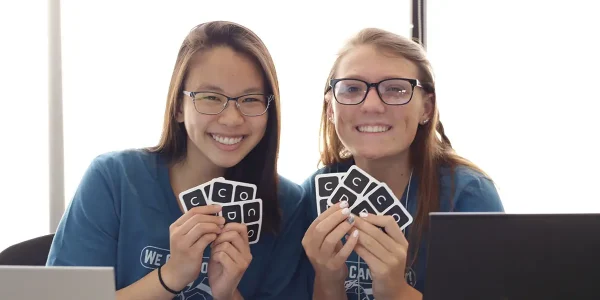 2 people sit at their laptops, each smiling and fanning out cards with "C-O-D-E" printed on them