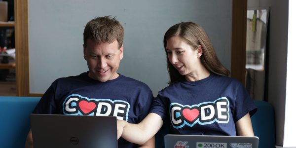 2 NewBoCo employees site behind their laptops and showcase their "Code" t-shirts with hearts replacing the "O's"