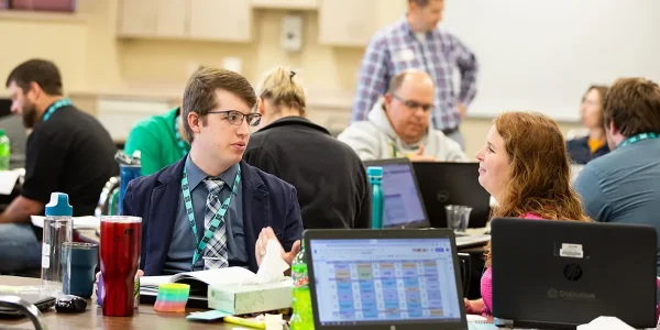 2 seated Code.org teachers hold a conversation, while more teachers in the background work with their heads down