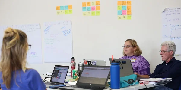 Teachers sit around their gathered laptops and engage in conversation; sticky notes cover the wall behind them